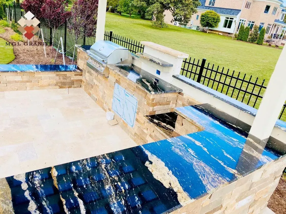 Outdoor kitchen with blue and gold countertop, stone structure, grill, and sink, overlooking a lawn.
