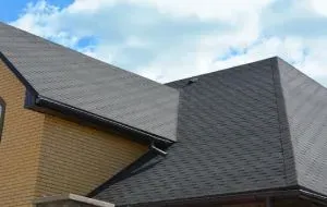 Beige brick house with gray shingle roof, dark gutters, and blue sky background
