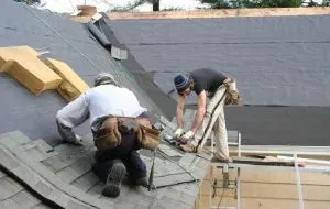Two roofers installing shingles on a sloped roof, one working with a tool, another kneeling, working