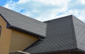 Beige brick house with gray shingle roof, dark gutters, and blue sky background