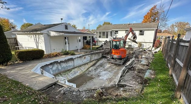 Backyard pool being excavated with an orange mini excavator beside a house and fence