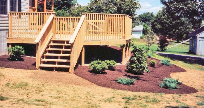 Wooden deck with stairs in a landscaped yard, featuring mulch beds and small shrubs