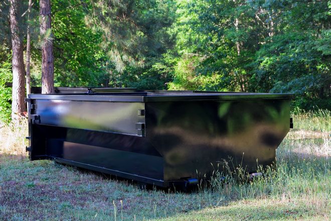 Black metal dumpster outdoors in a grassy, wooded area