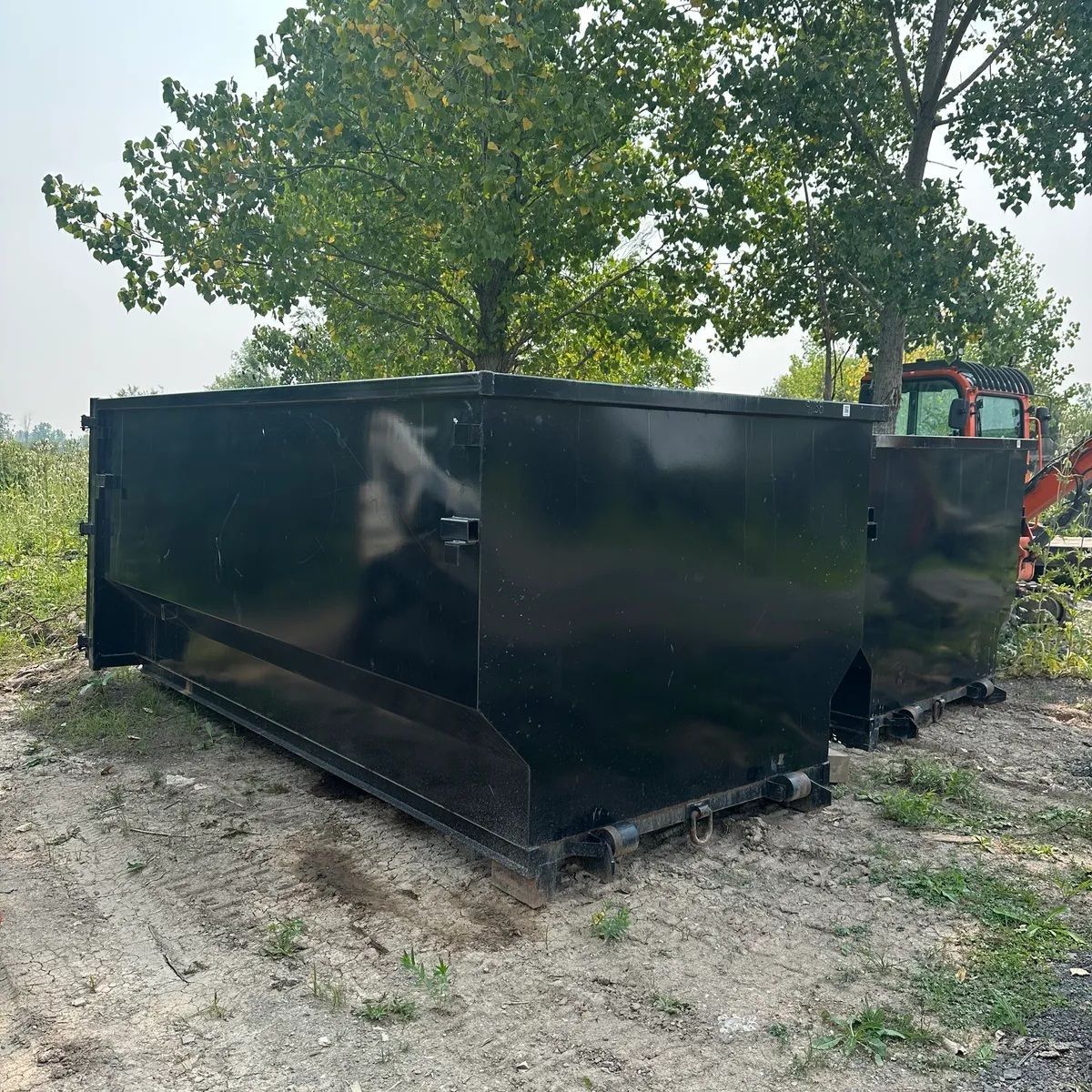 Black metal dumpster on dirt lot with a tractor behind it and trees in the background