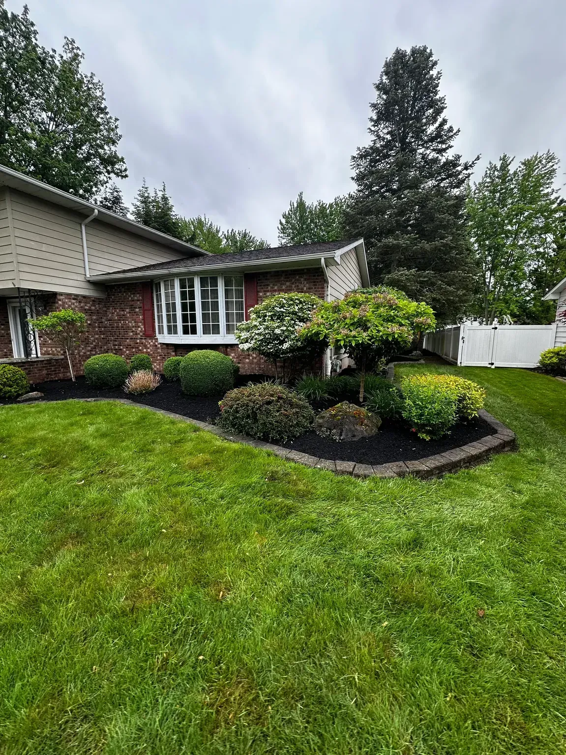 Front yard with green lawn, brick house, shrubs, and a white fence under cloudy sky