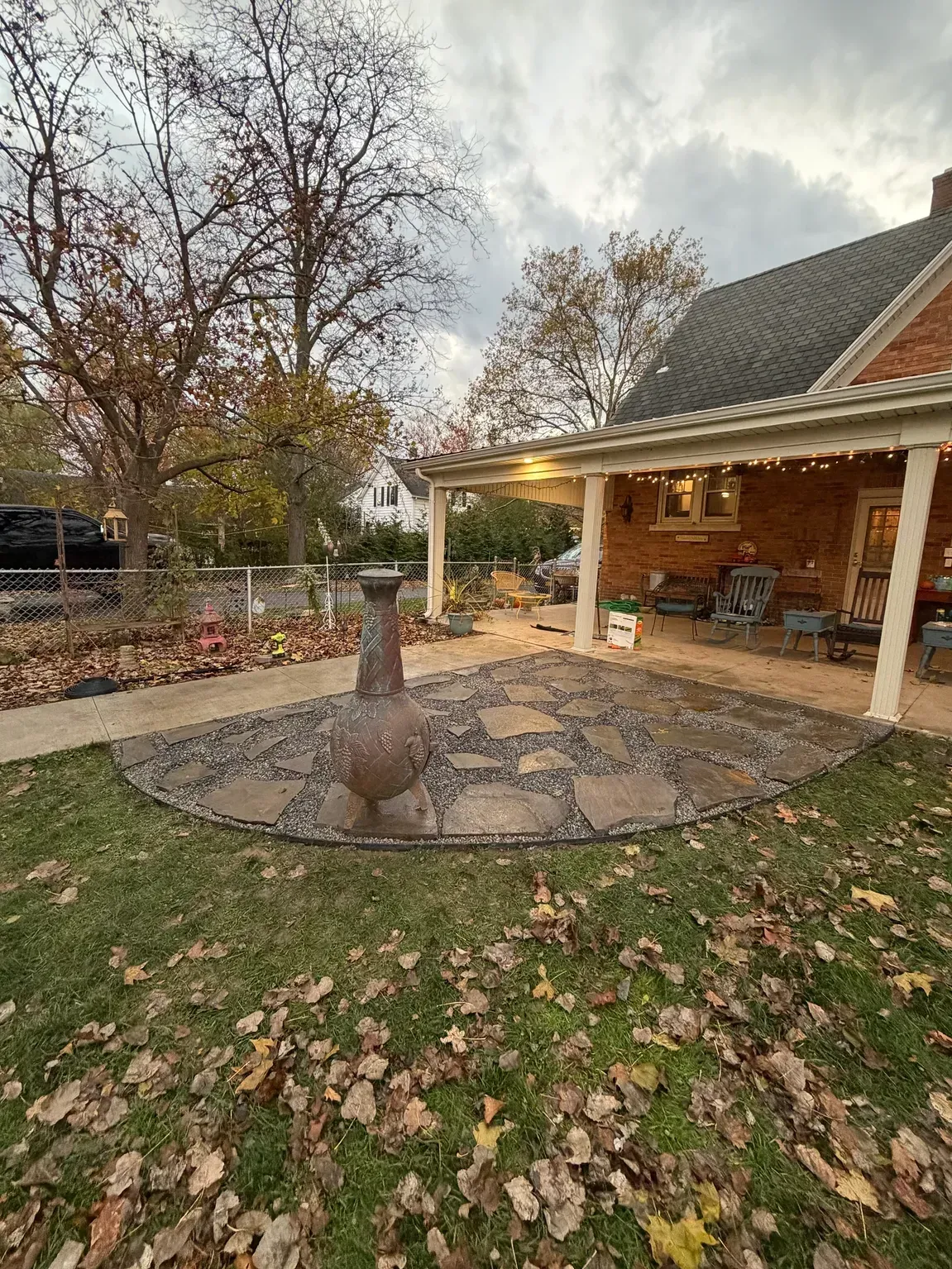 Front yard with circular stone patio, central fire pit, and covered porch beside autumn trees and fallen leaves