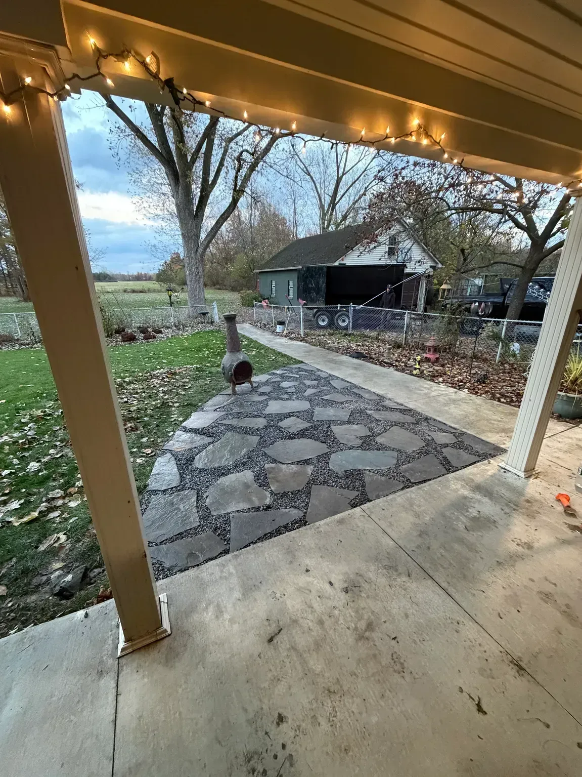 Covered porch overlooking stone patio, leafless trees, and a backyard shed at dusk