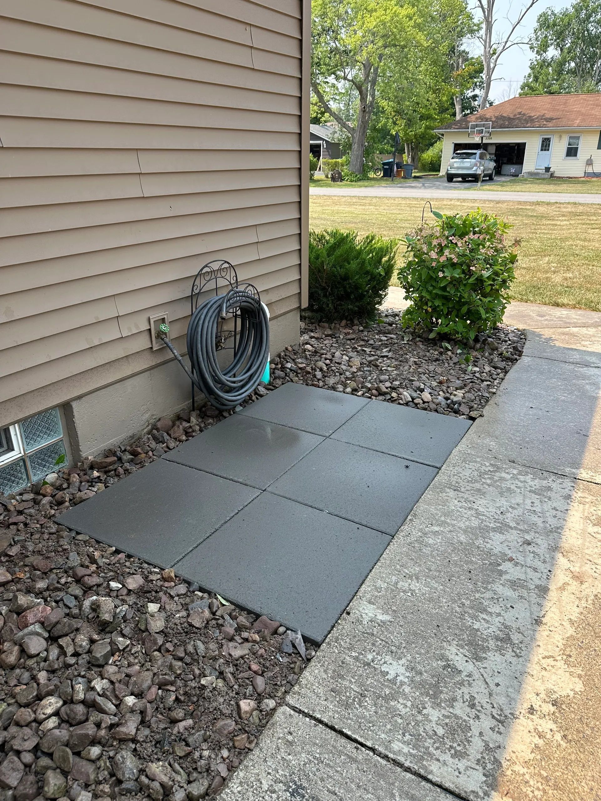 Side of house with coiled hose, concrete pavers, rocks, and bushes beside a sidewalk.