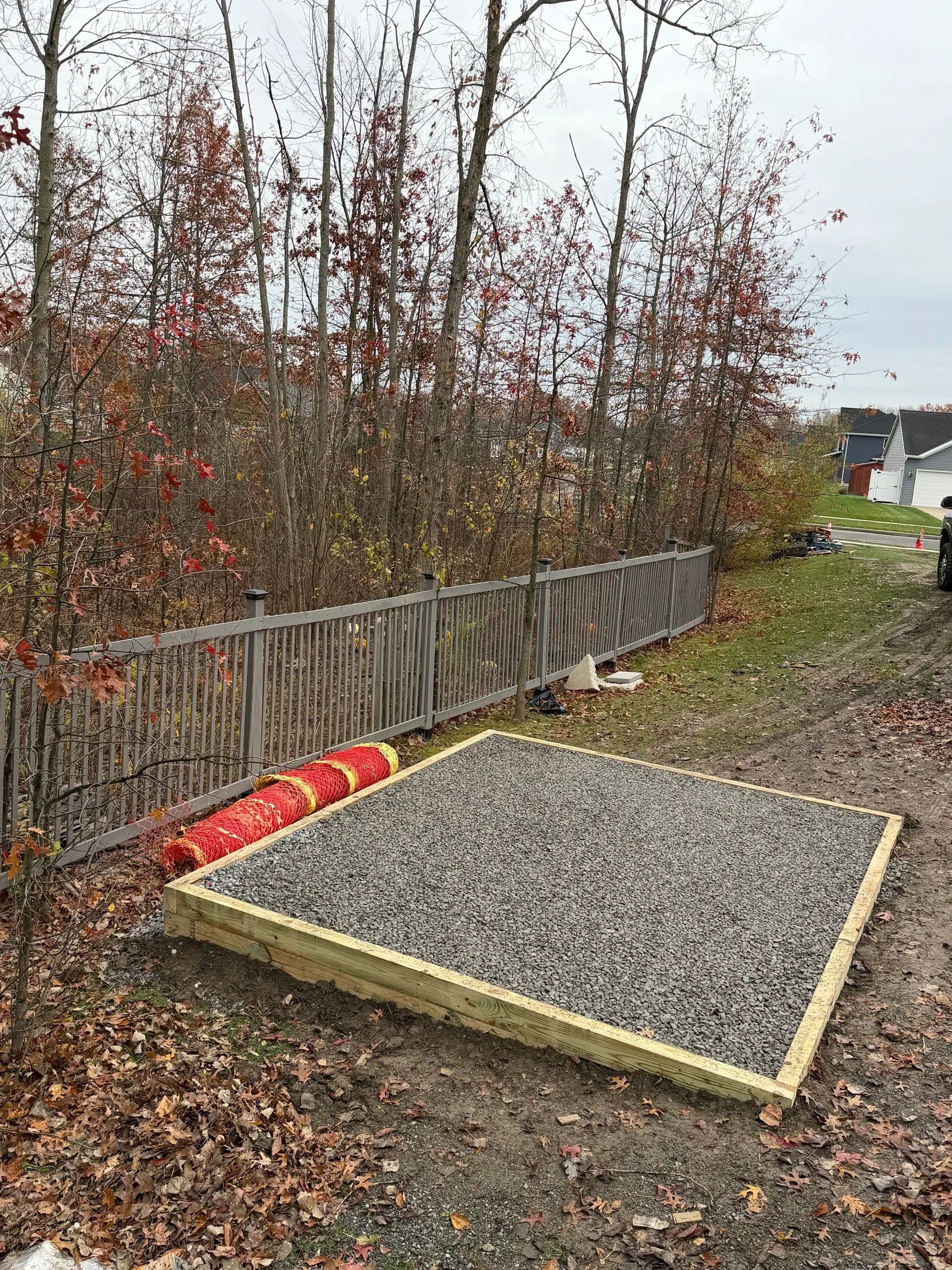 Gravel-filled wooden frame beside a fence and leafless bushes, with red pipes and scattered autumn leaves.