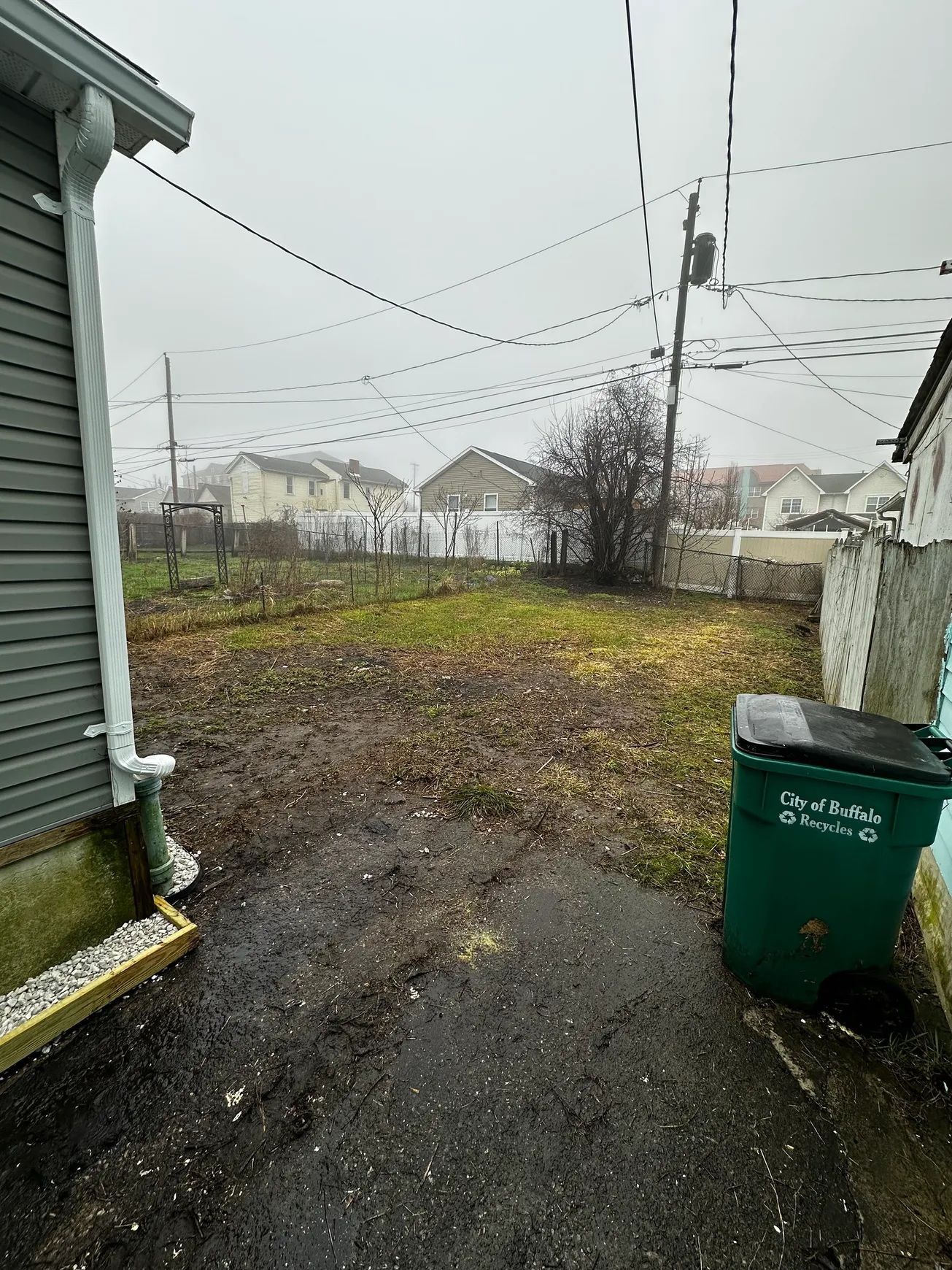 Rainy backyard beside a house, with a wet driveway, green trash bin, and snowy houses in the distance