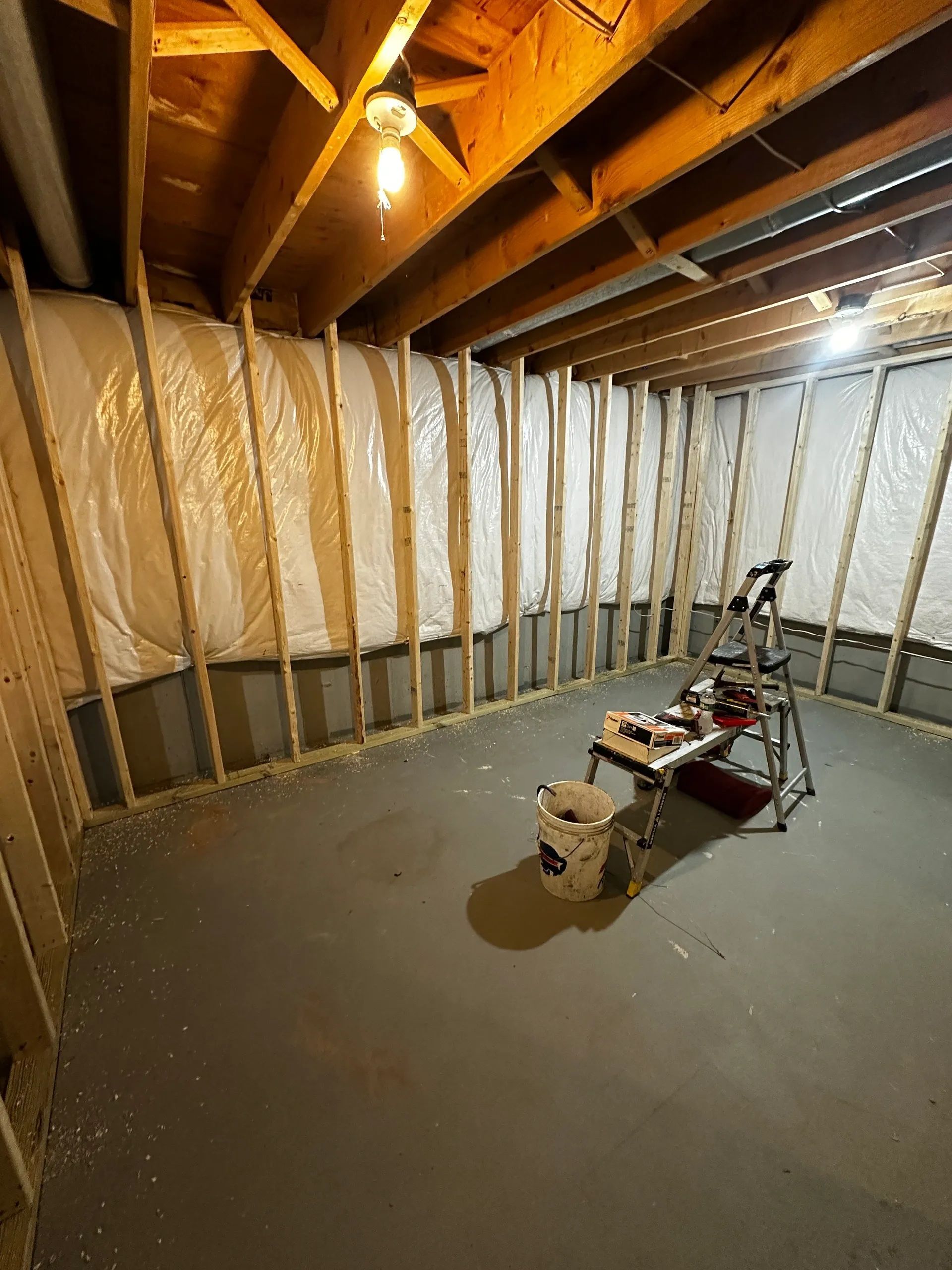 Basement under construction with exposed joists, insulation, and tools on a concrete floor.