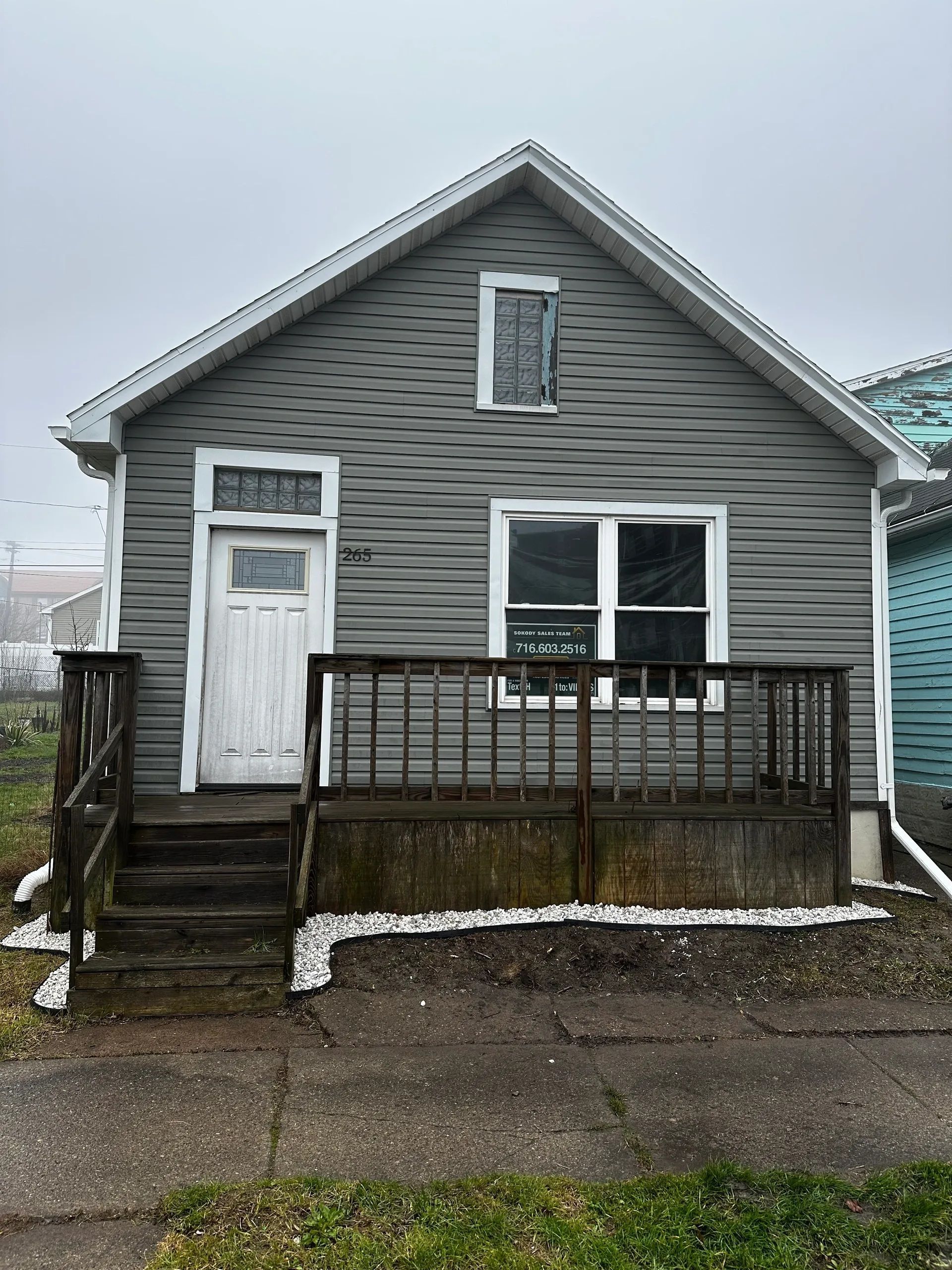 Small gray house with a white door, front porch, and steps on a wet street