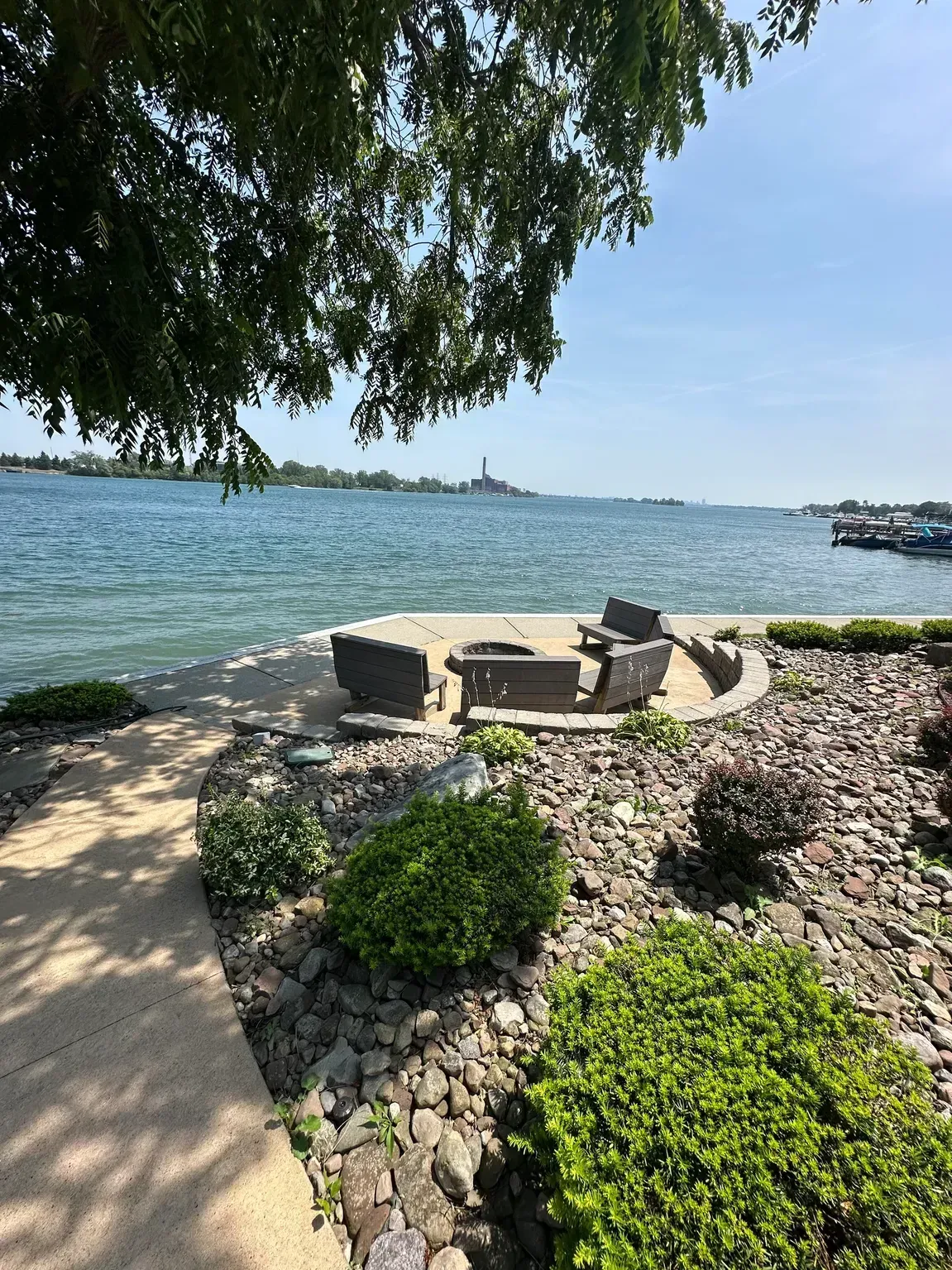 Lakefront patio with stone seating, rocks, and greenery under a tree beside blue water and a distant skyline