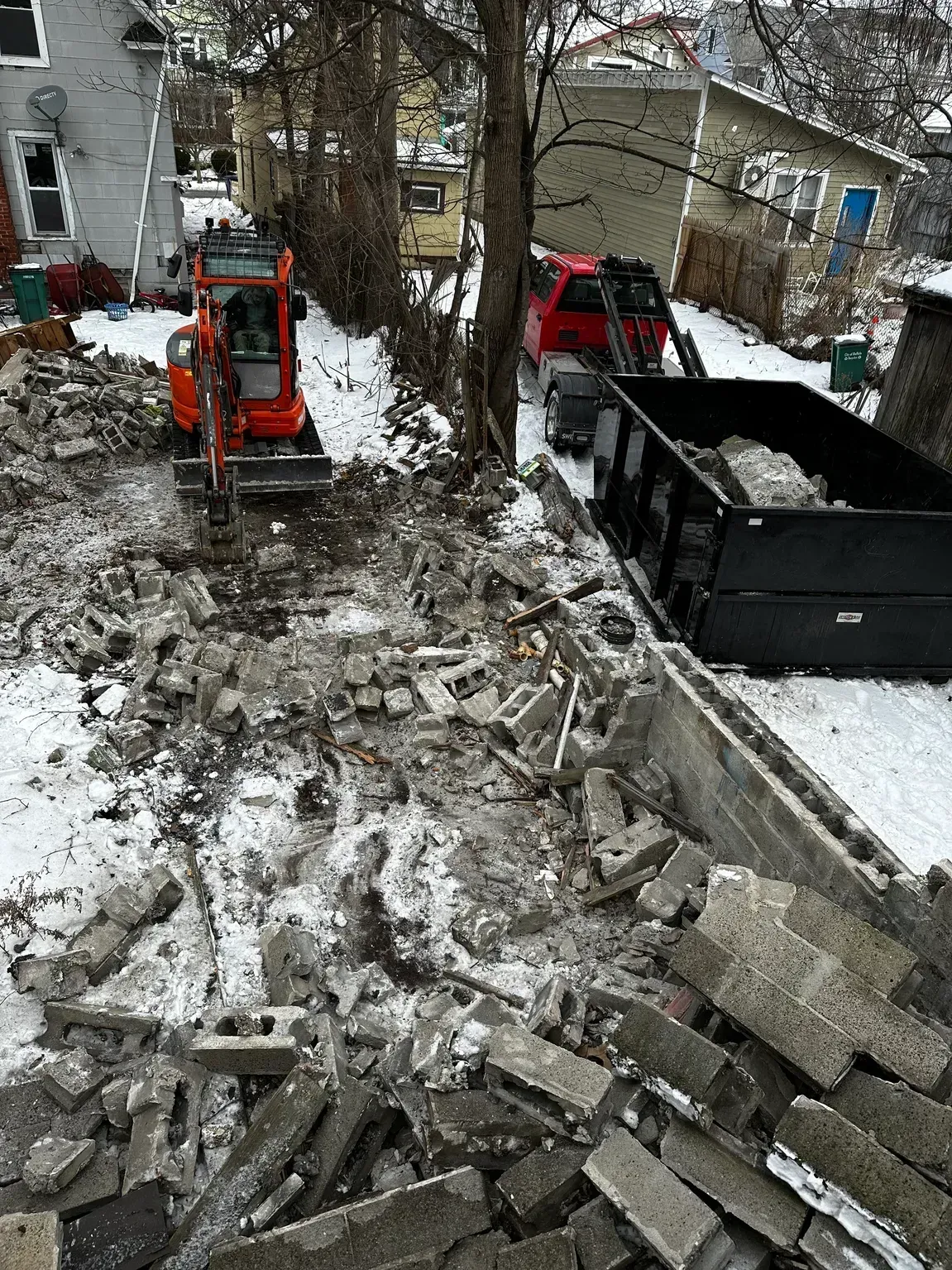 Construction site with excavator and dump truck clearing debris in a snowy yard