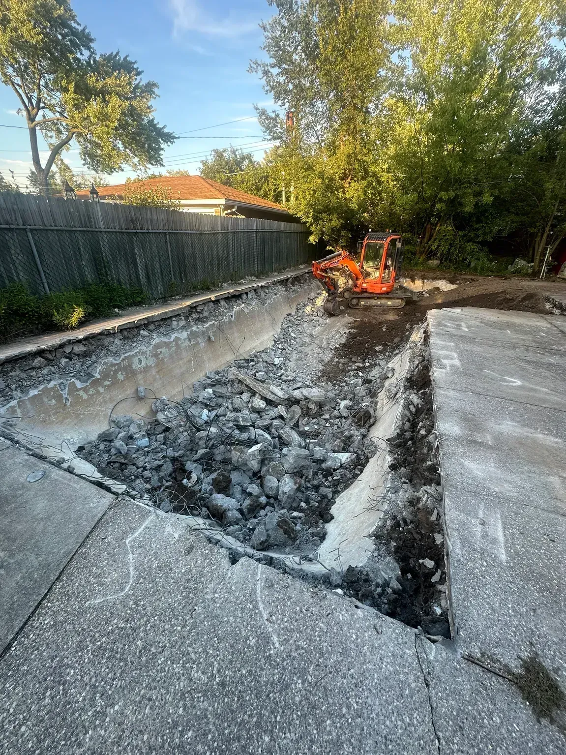 Excavator digging up a cracked concrete driveway beside a fence and trees outdoors