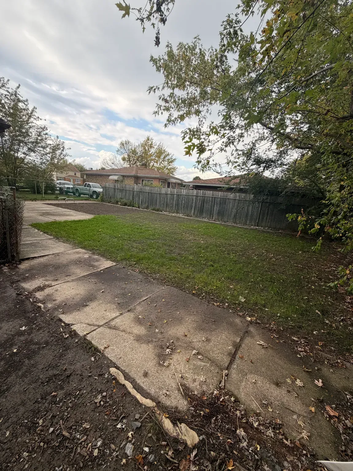 Cracked concrete path beside grassy yard, trees, and a stone wall under a cloudy sky