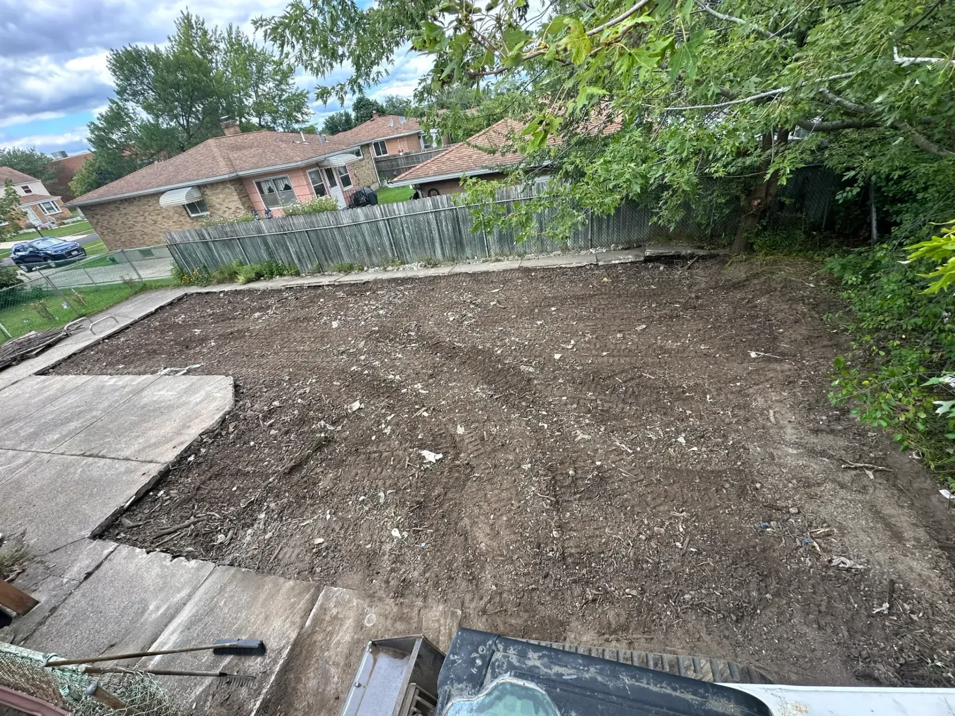 Freshly leveled dirt lot beside a stone wall, with houses and trees in the background