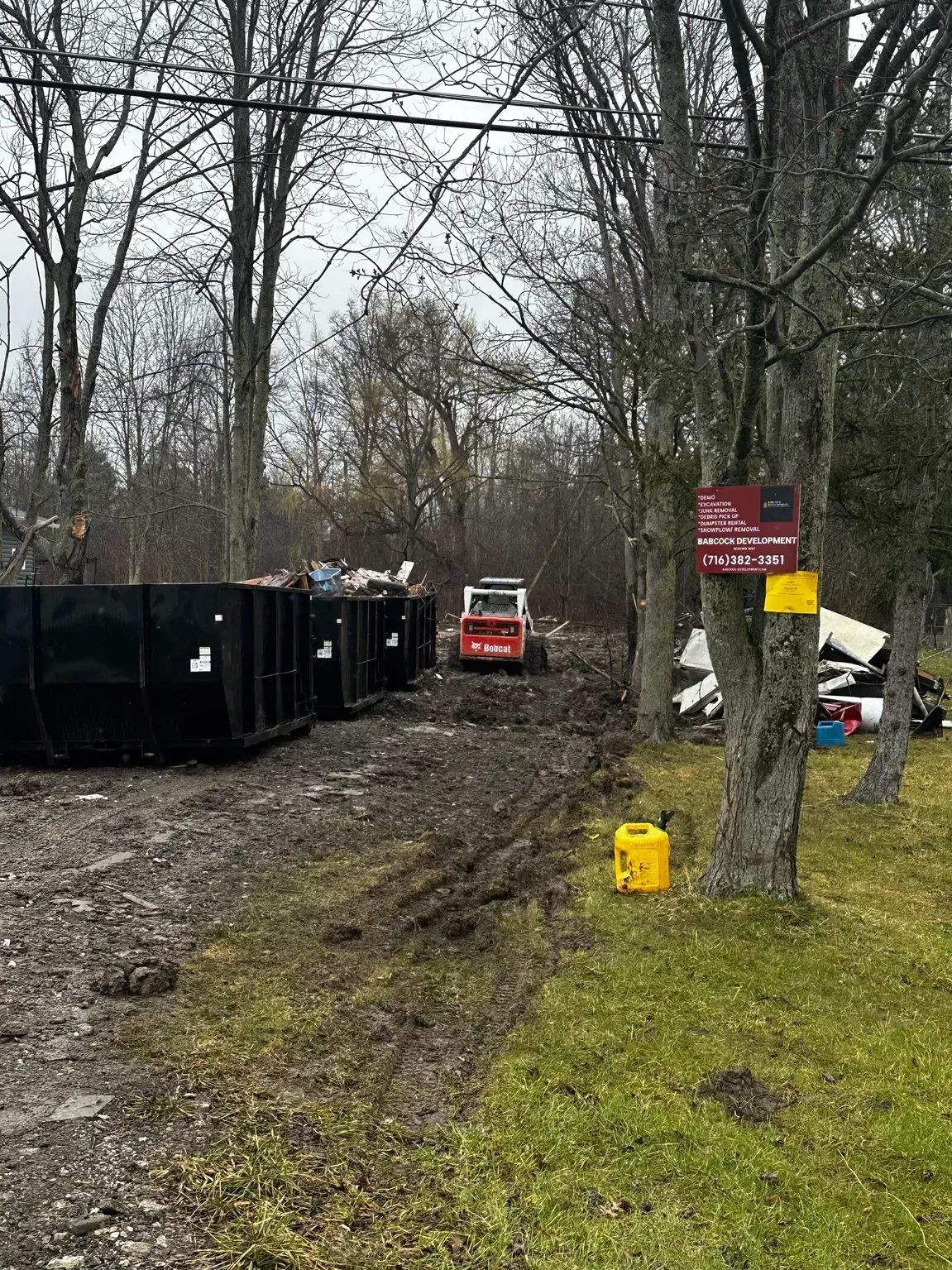 Red skid-steer near black dumpsters on a muddy wooded lot, with a yellow sign on a tree.