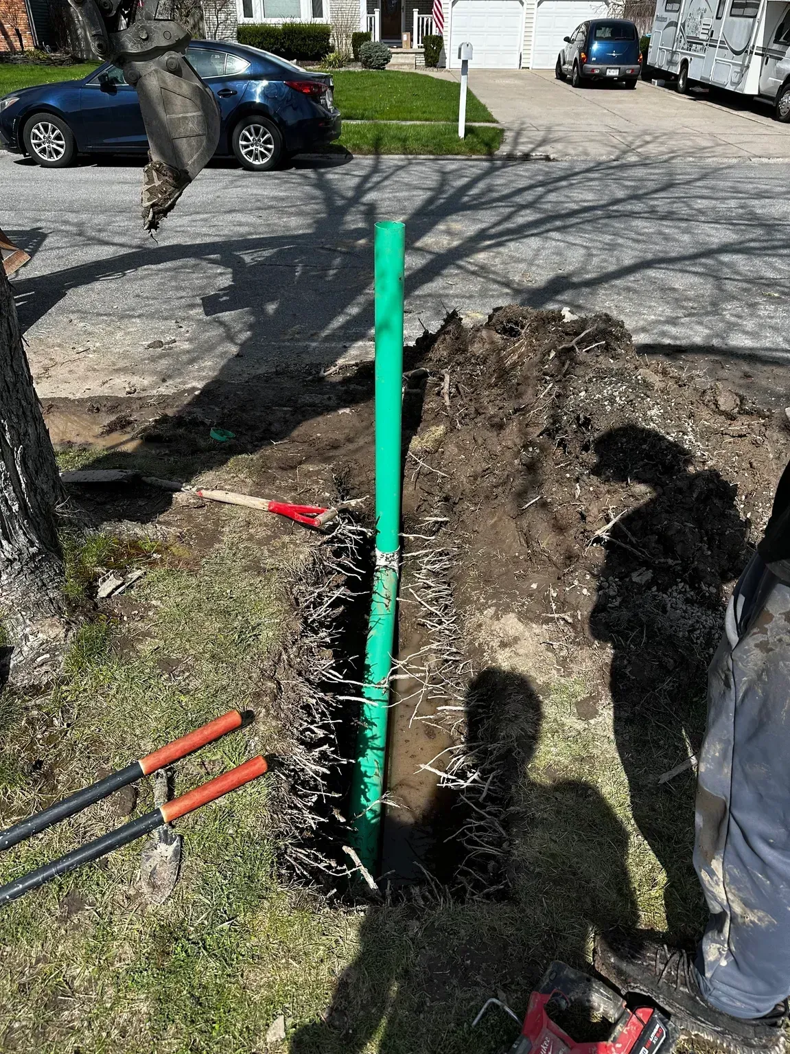 Green utility marker post beside an open trench in a yard, with tools and workers nearby.