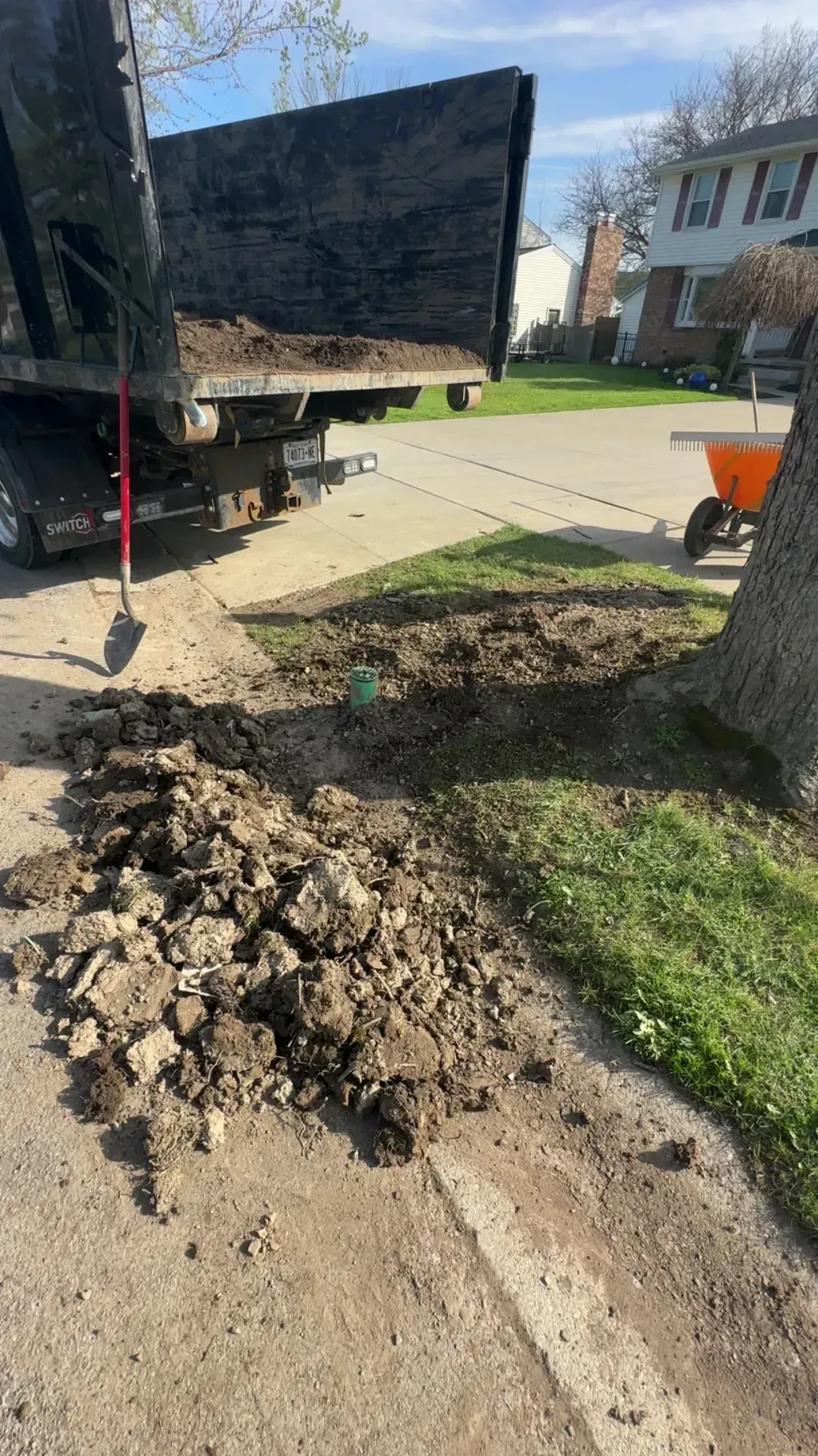 Backyard sidewalk with muddy dirt piles beside a large black truck and a tree, near a house.