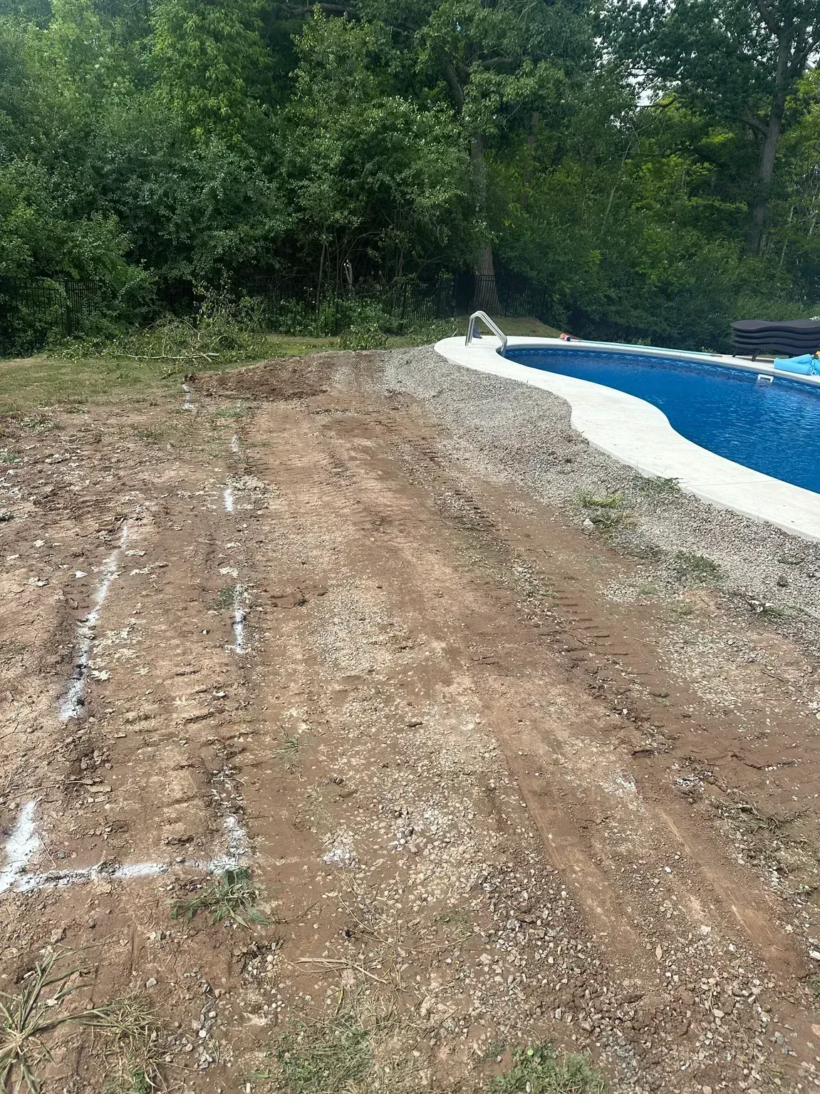 Bare dirt path beside a blue swimming pool, with trees and brush in the background.
