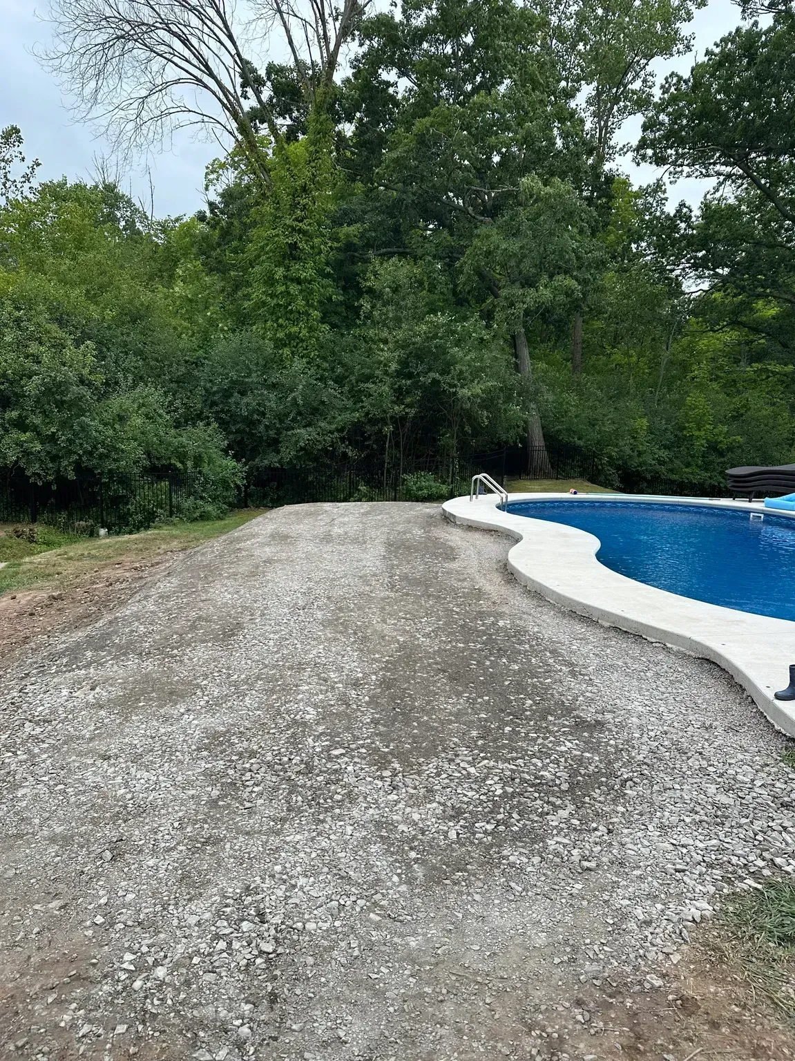 Gravel path beside a blue swimming pool, surrounded by dense green trees and bushes.