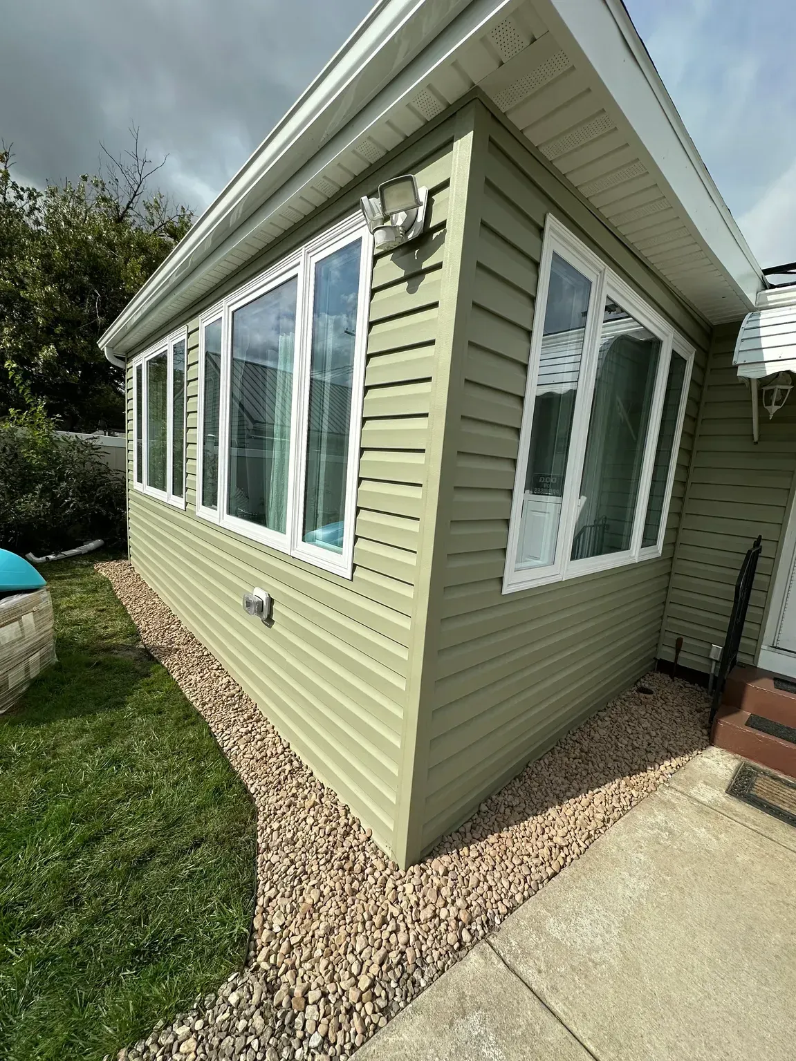 Small beige house exterior with large windows, white trim, and a gravel border beside a concrete walkway.