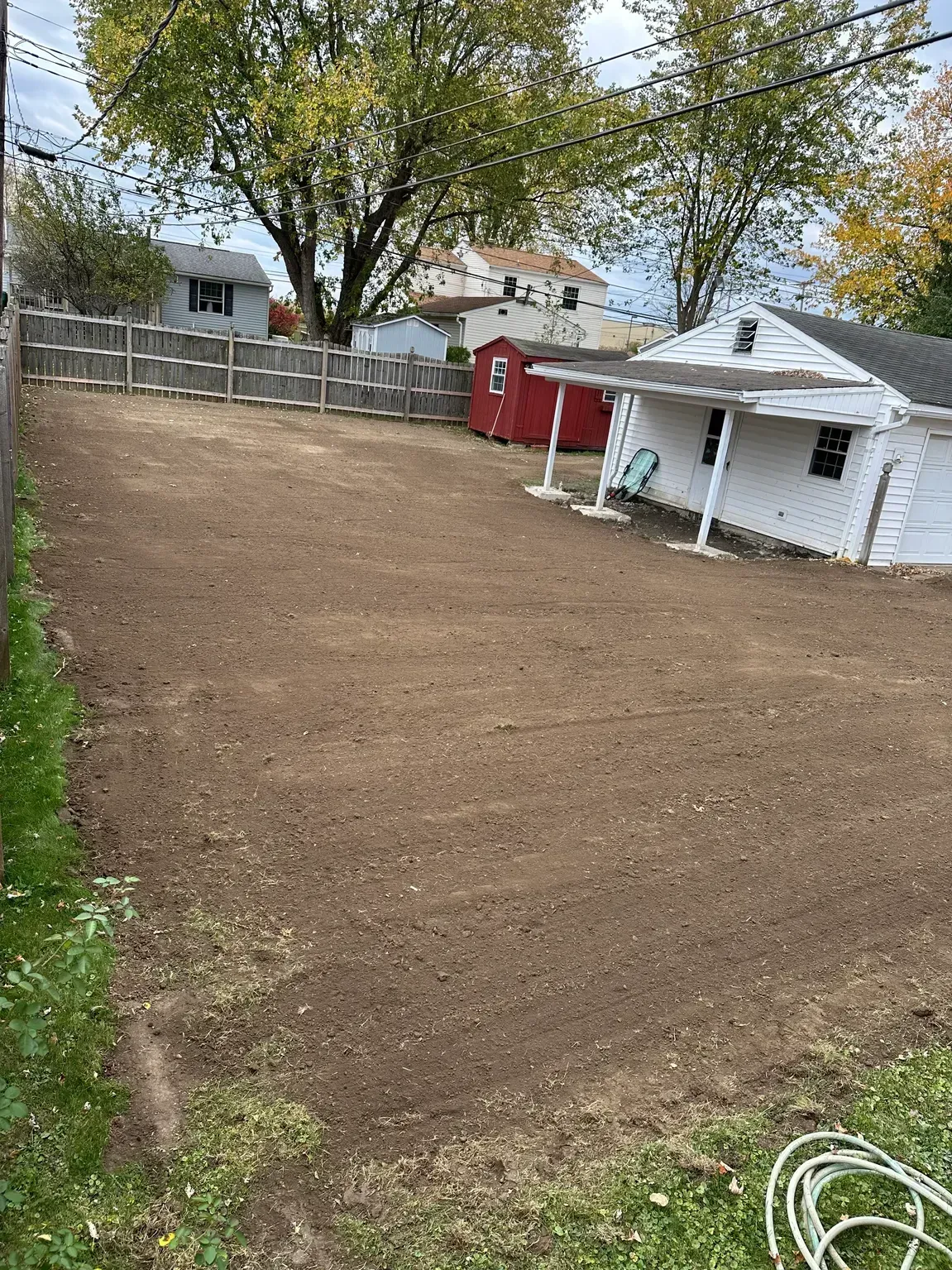 Freshly tilled backyard with a white shed, red gate, and trees in autumn.