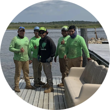 Five people in green shirts stand on a dock by the water, with a couch in the foreground.