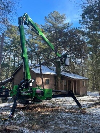 Green spider lift with a worker raised beside a cabin in a snowy pine forest.