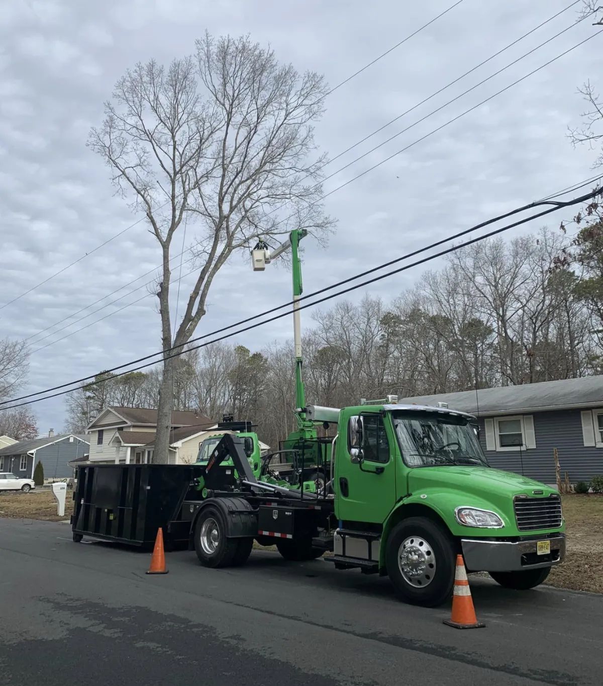 Green utility truck with lift parked on a residential street beside cones and a large black dumpster