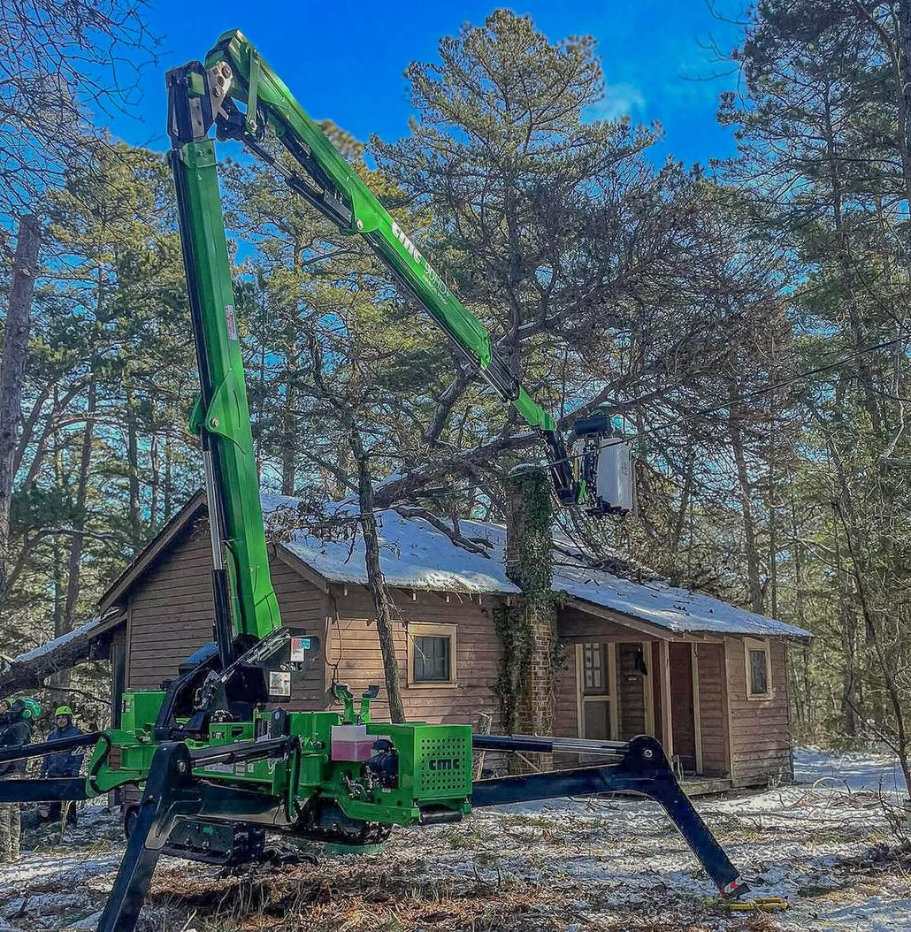 Green forestry machine lifting a worker beside a small log cabin in a wooded area