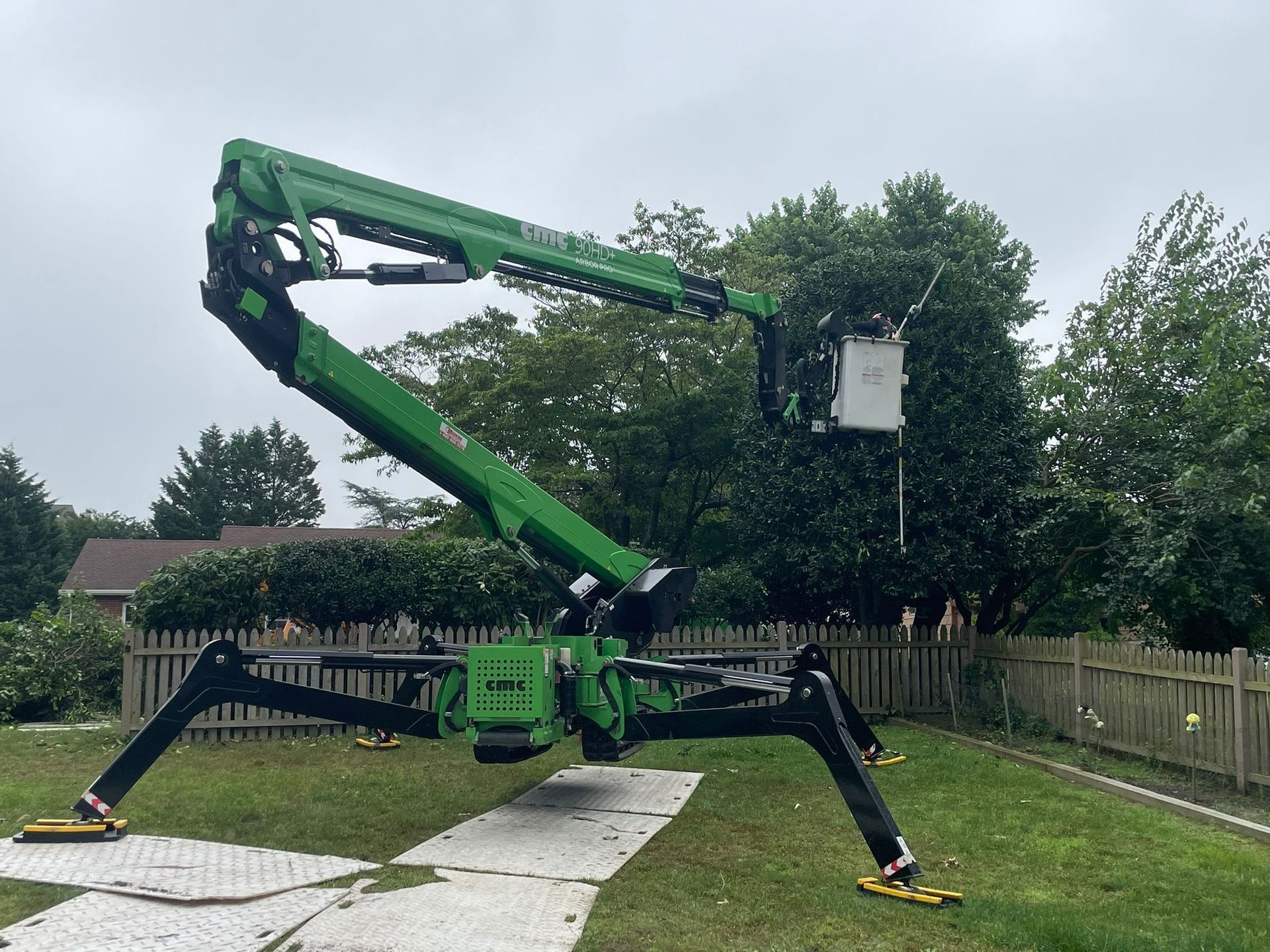 Green spider lift parked on a driveway beside a fence and trees, with outriggers extended.