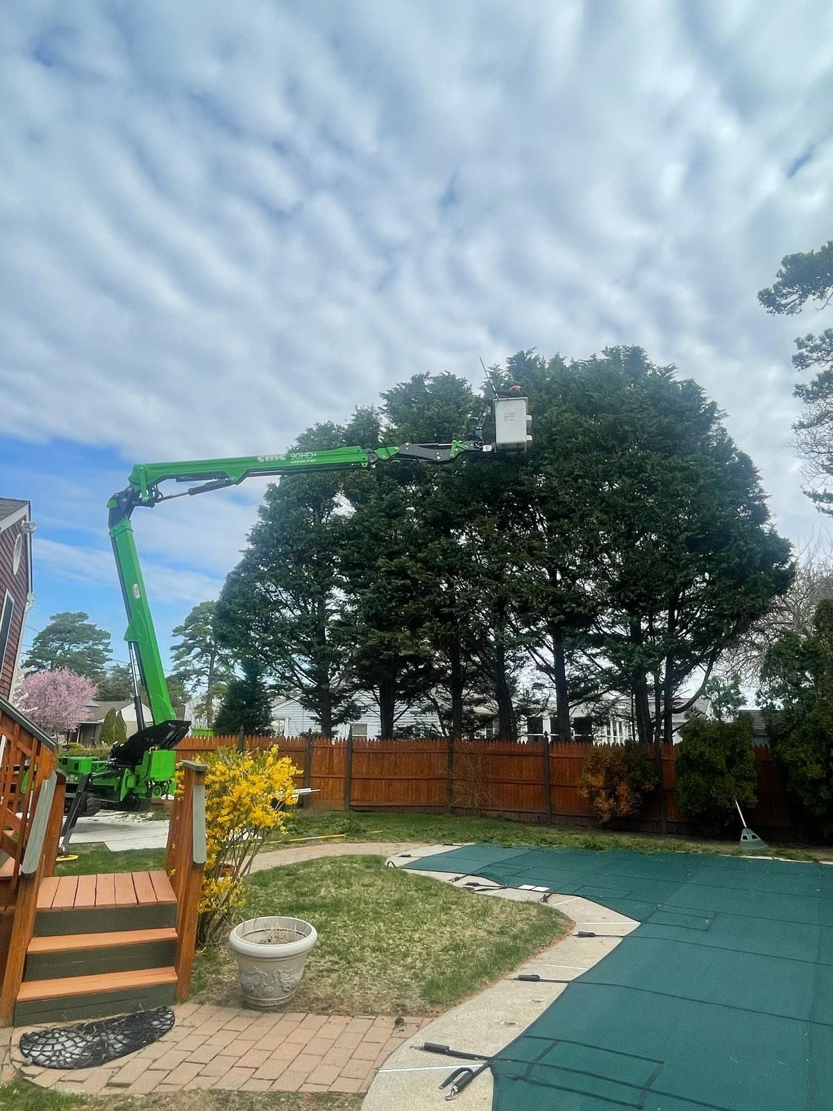 Backyard with a green boom lift beside a wooden fence, patio, lawn, and large trees under a blue sky