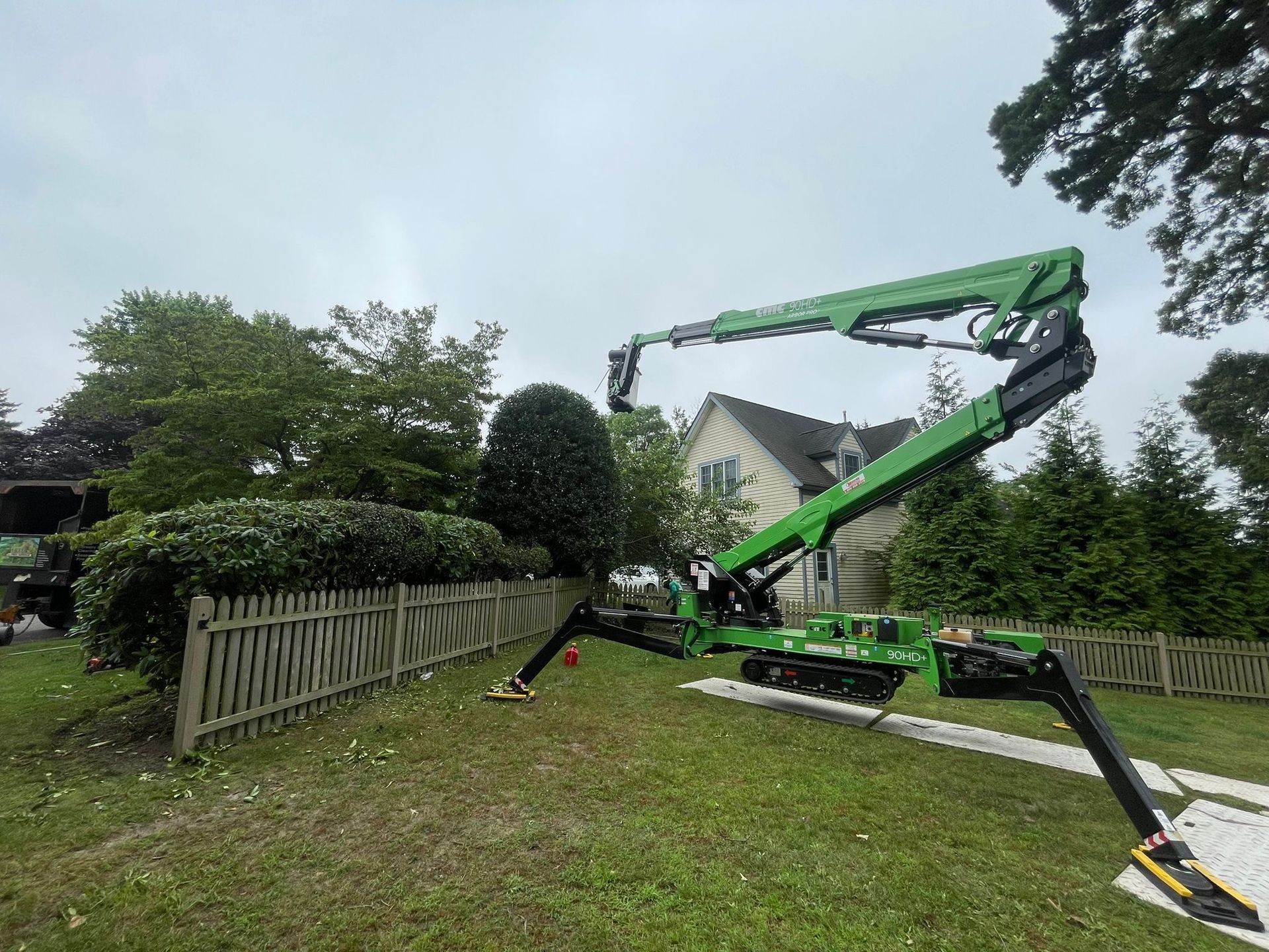 Green crane lifting a fallen tree beside a white fence in a grassy yard