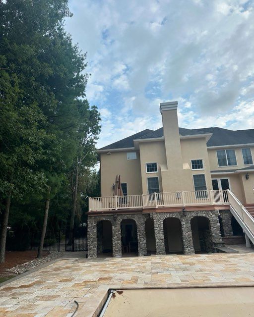 Large beige two-story house with stone arches, a tall chimney, and a paved driveway beside trees.