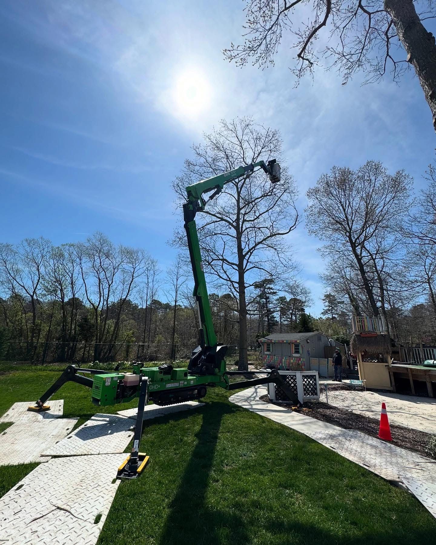Green tree-trimming lift in a sunny yard with workers' equipment and a house in the background