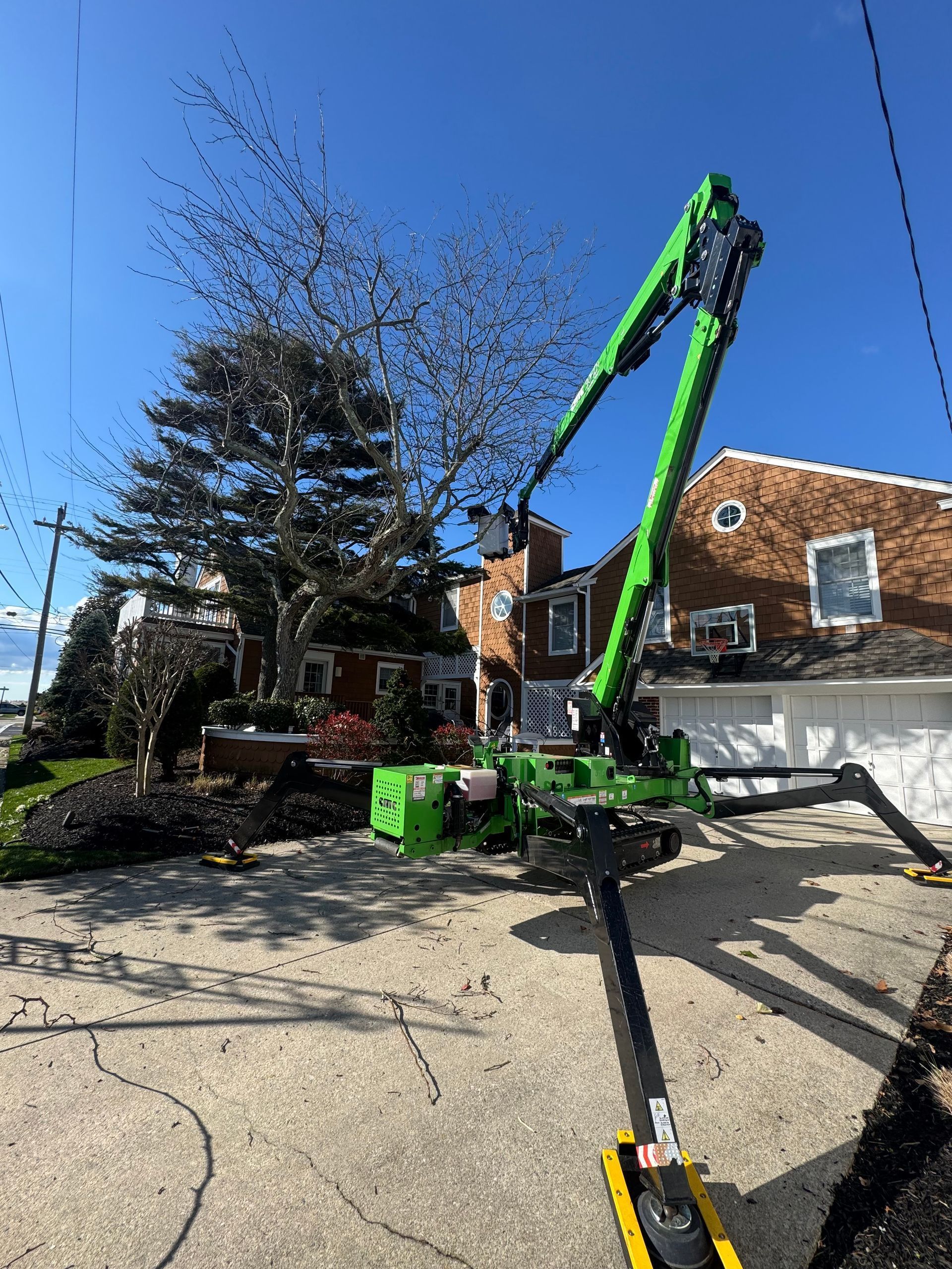 Green boom lift extended beside a brick house on a sunny driveway, with a tree and blue sky.