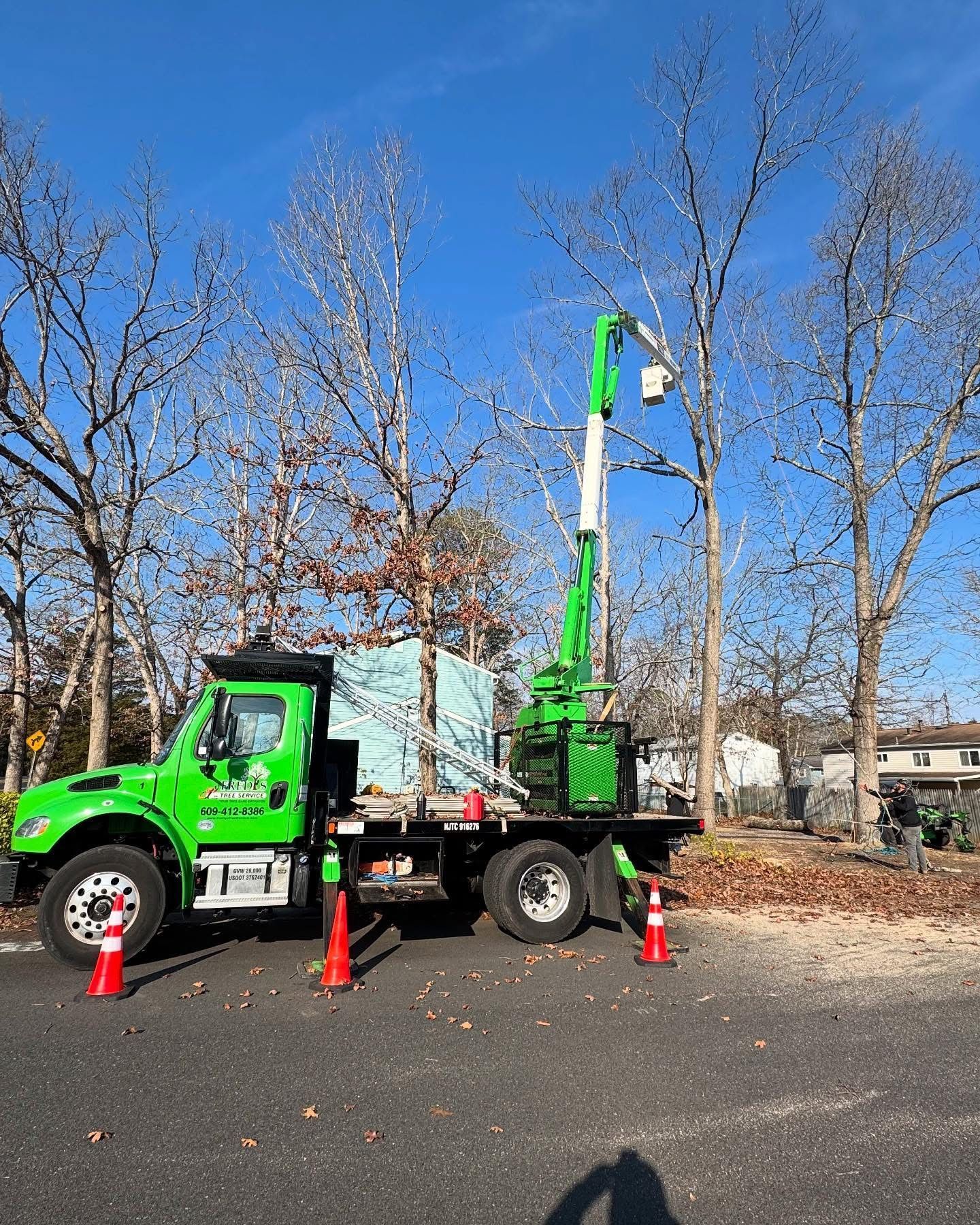 Green utility truck with raised boom parked by a roadside, with orange cones and leafless trees nearby