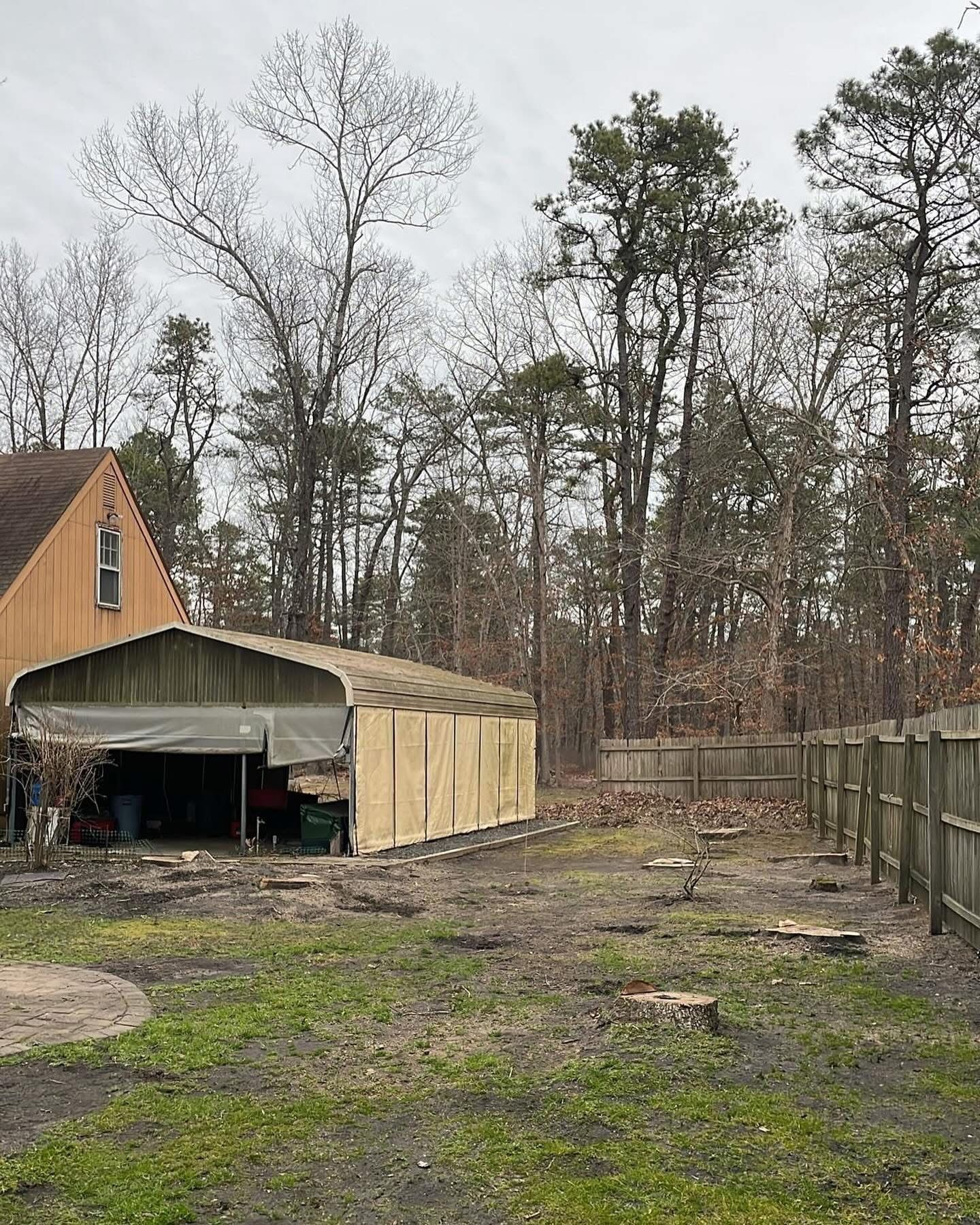 Muddy backyard beside a tan shed and house, with a wooden fence and leafless trees in the background