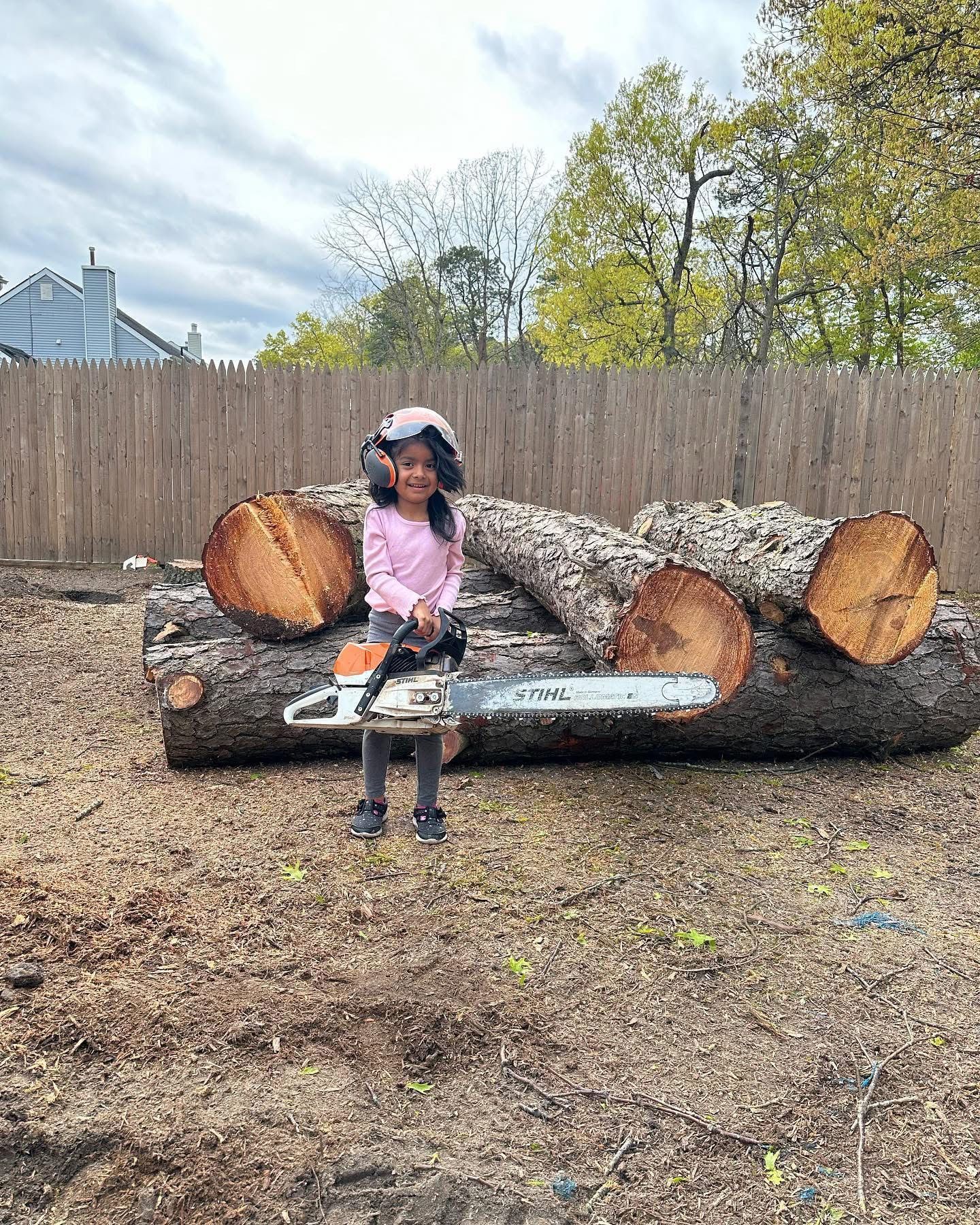Child in pink jacket seated on a large fallen tree trunk in a yard, with stacked logs and trees behind.