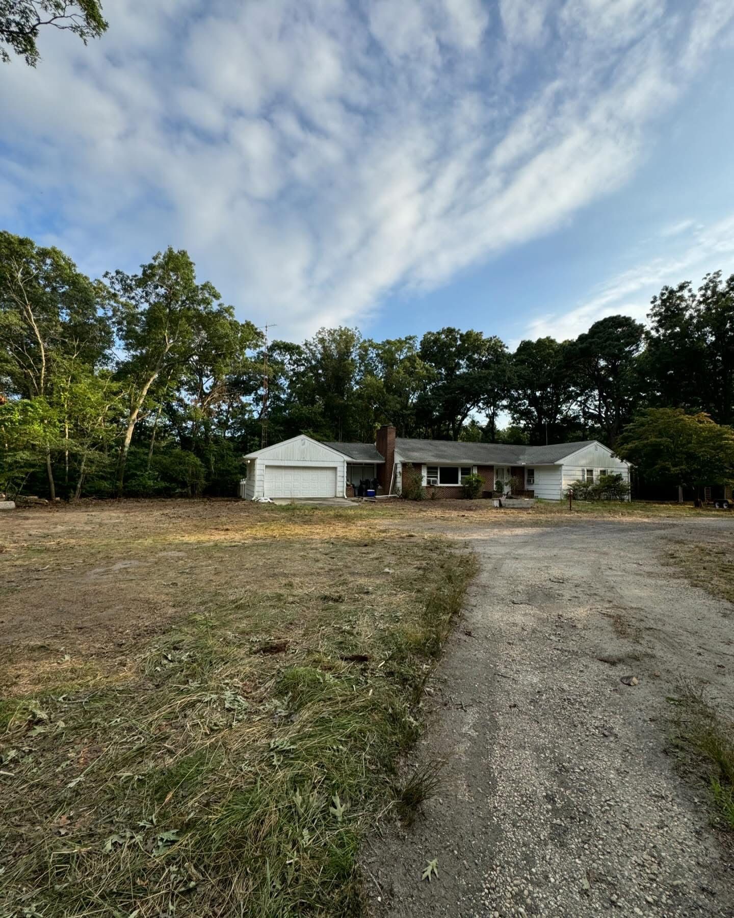 Rural house with white outbuildings, dirt driveway, and trees under a partly cloudy sky