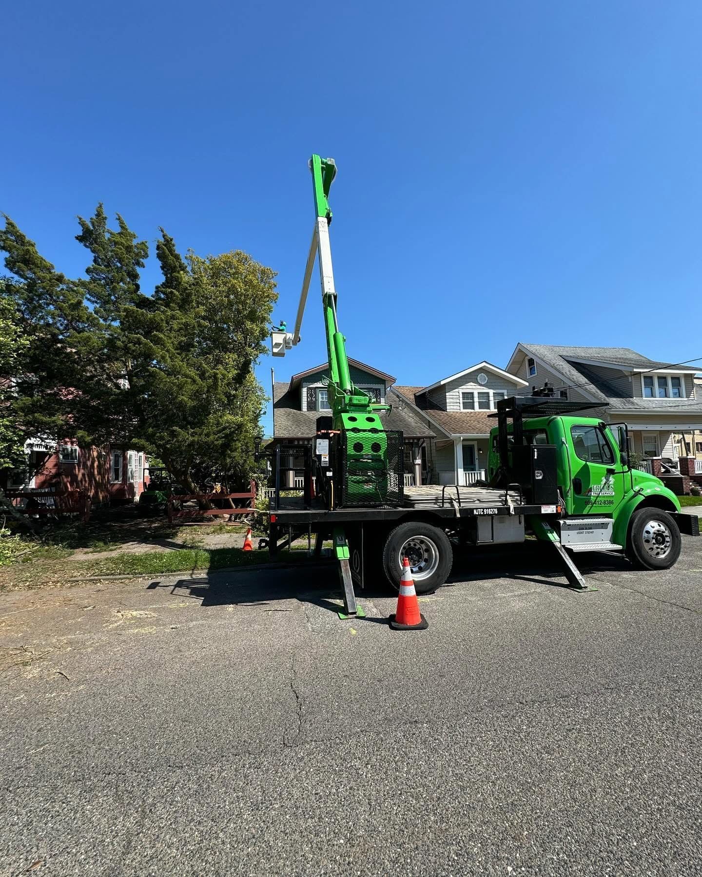 Green utility truck with lift parked on a driveway beside a house, with an orange cone nearby.