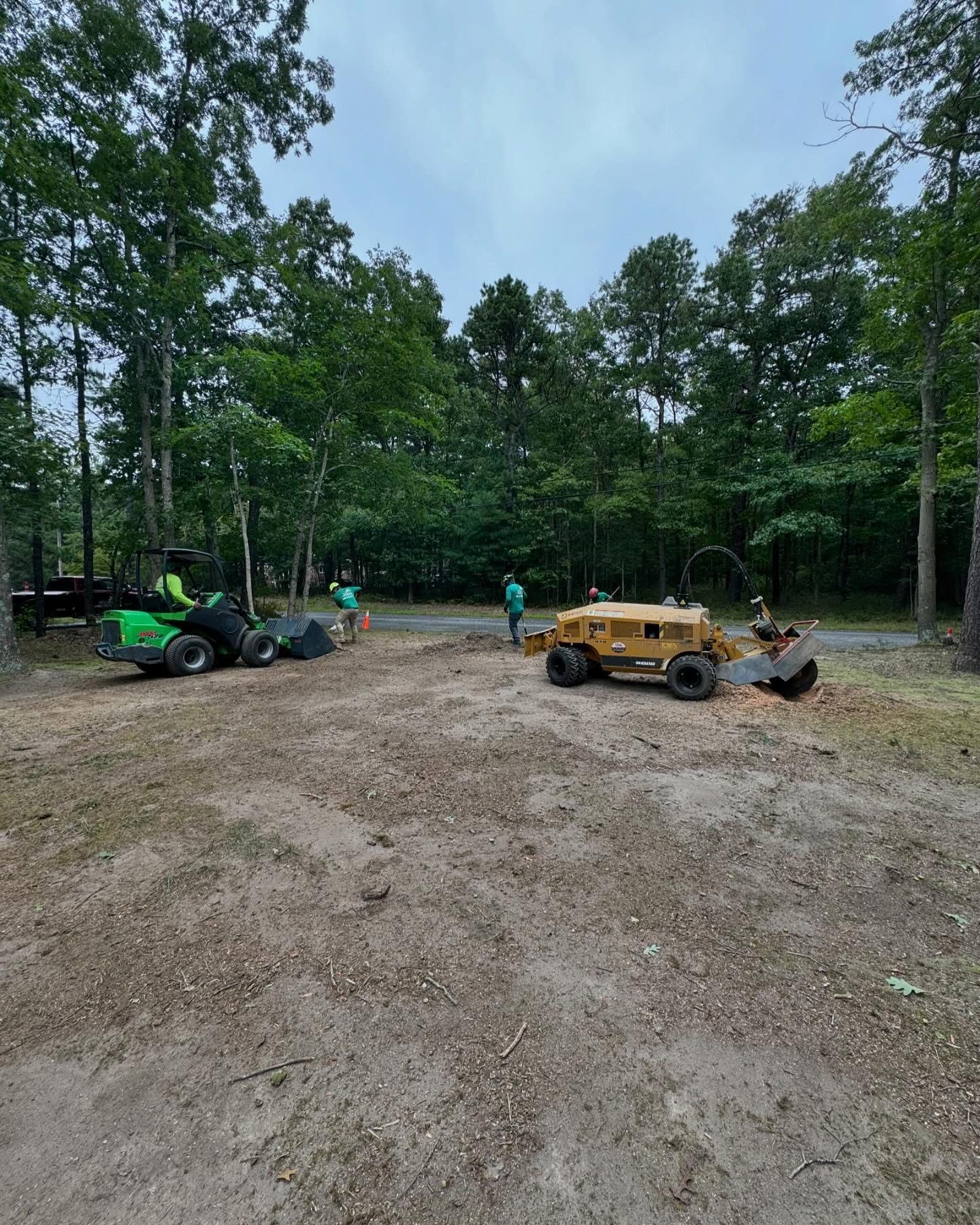 Two mini excavators, green and yellow, on a dirt lot beside a wooded tree line