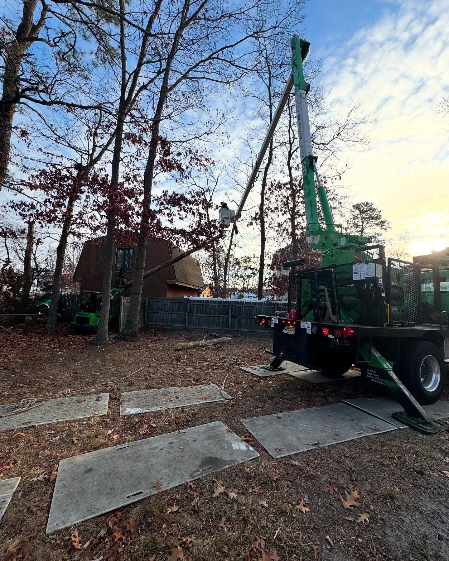 Green crane truck lifting a tall pole beside a house on a leaf-covered yard at dusk