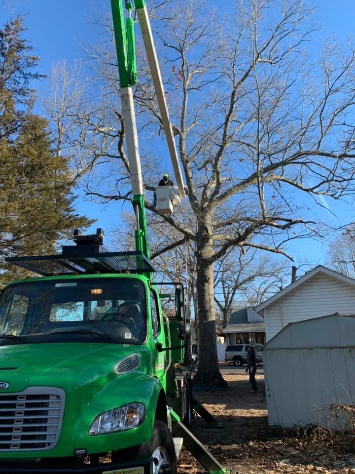 Green utility truck with raised boom parked beside a house and leafless tree under a blue sky