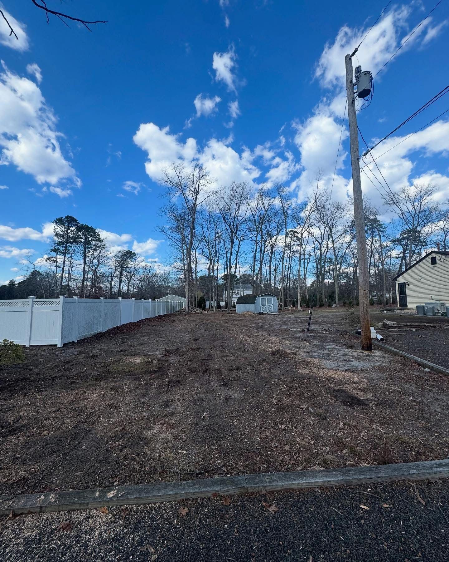 Vacant dirt lot beside a white fence and utility pole under a blue sky with clouds
