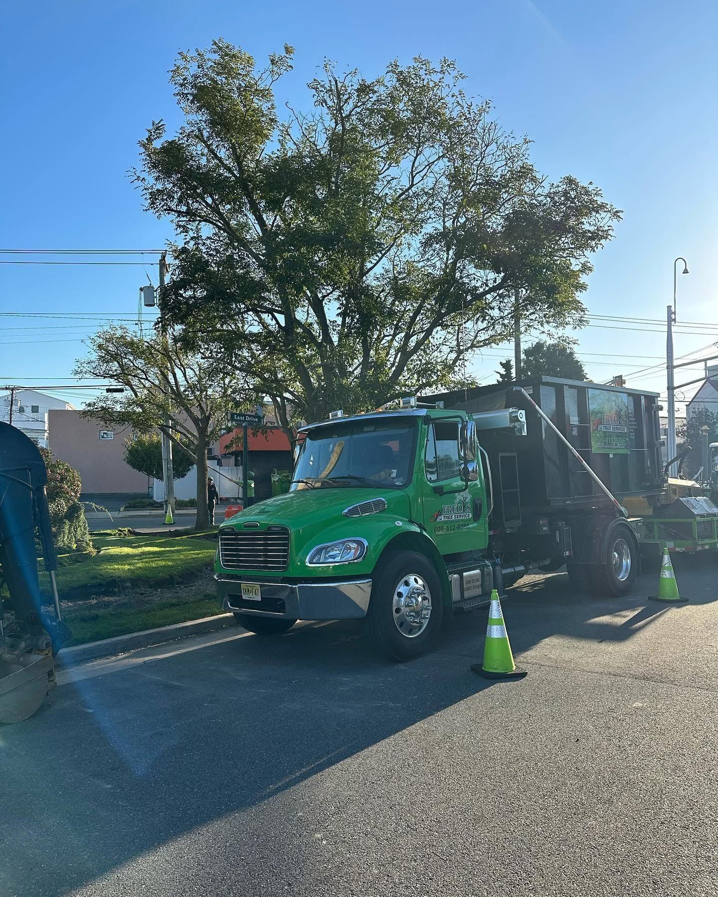 Green tow truck parked on roadside with cones, under a tree in bright sunlight.