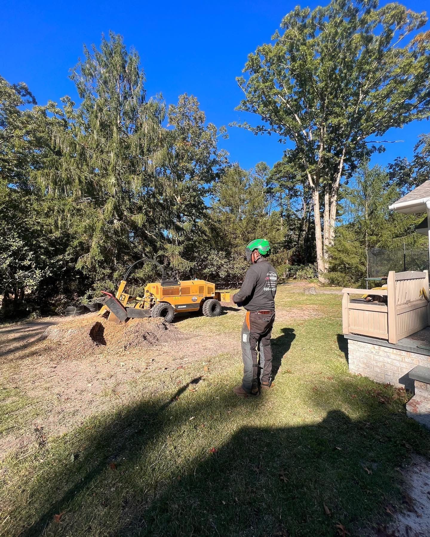 Person in a green hard hat stands in a sunny yard beside a small yellow excavator and dirt pile.