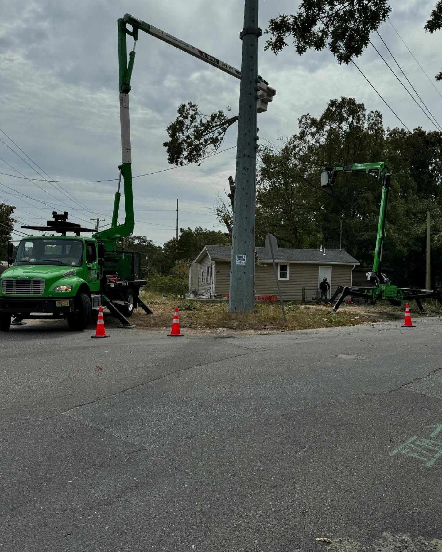 Utility crews using green boom trucks work on power lines beside a small house, with cones blocking the road.